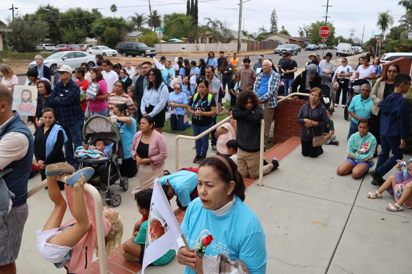 Crowd gathered on a sidewalk, with people holding signs and a stroller near a red stop sign.