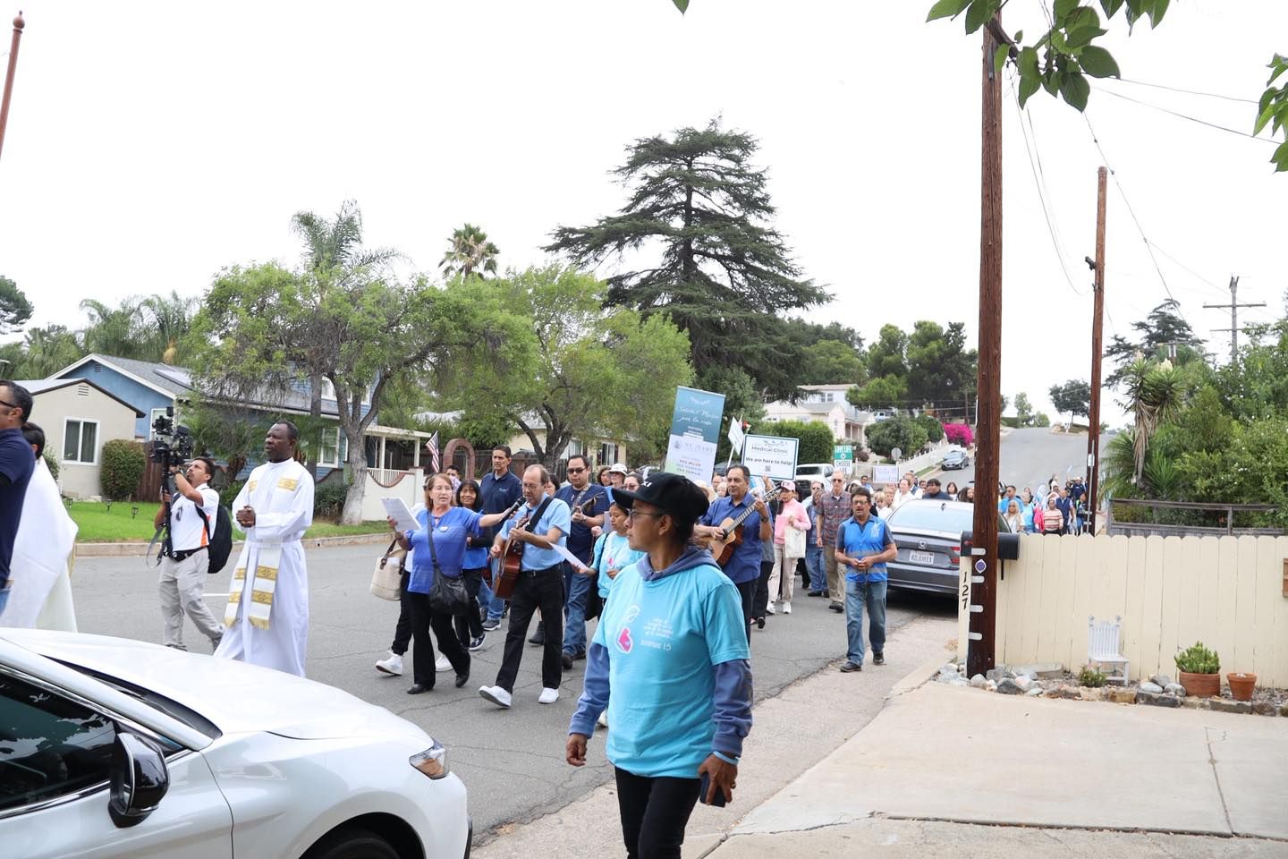 Crowd marching in a street procession, with people in blue and white clothing near parked cars and trees.
