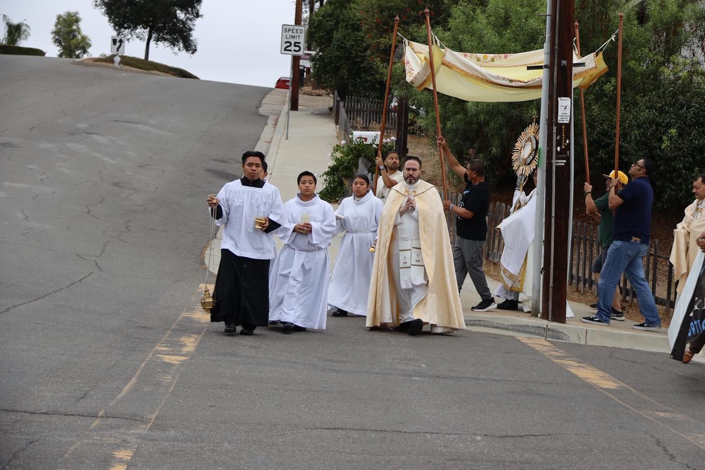 Clergy procession walking down a steep road under a canopy, with onlookers nearby.