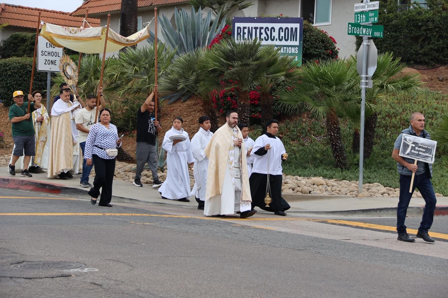 Priests and altar servers in white robes walk in a street procession past spectators and homes.