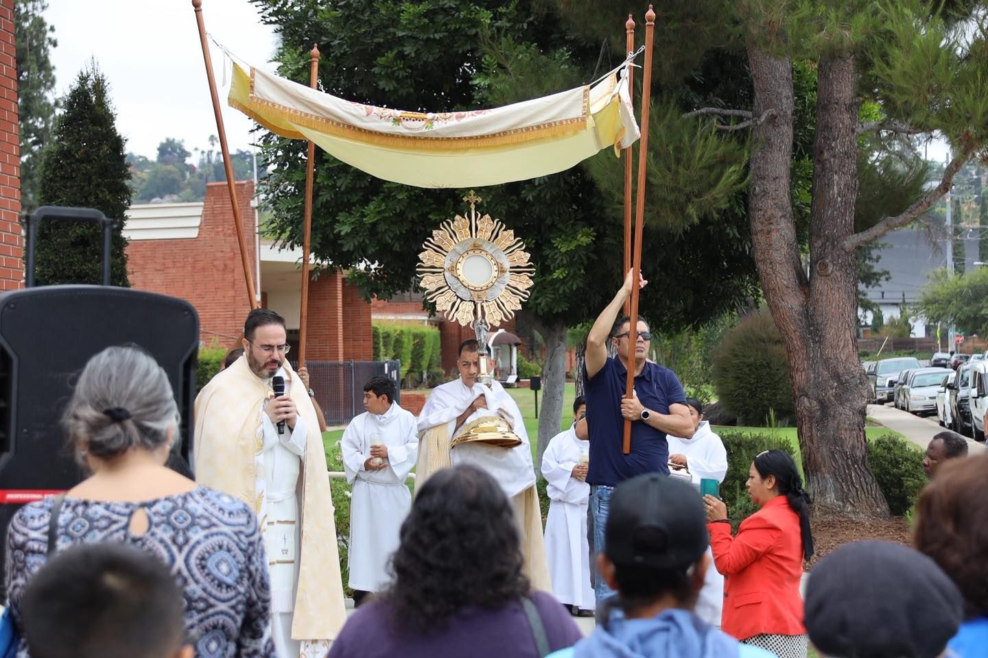 Outdoor Catholic procession with a priest holding a monstrance under a canopy, surrounded by worshippers