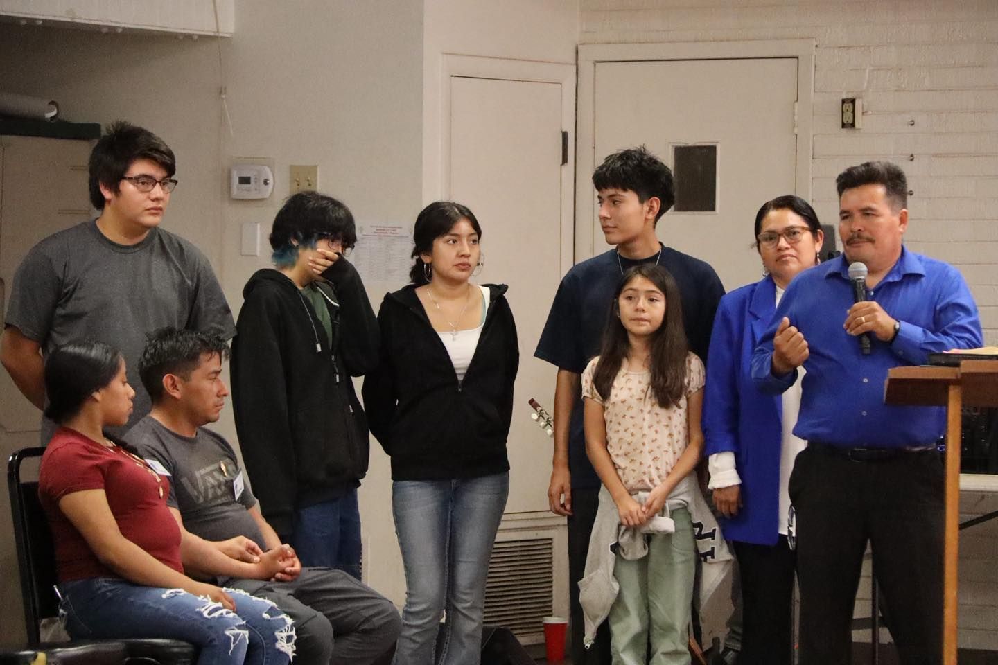 Several people stand and sit in a room as a man in a blue shirt speaks into a microphone.
