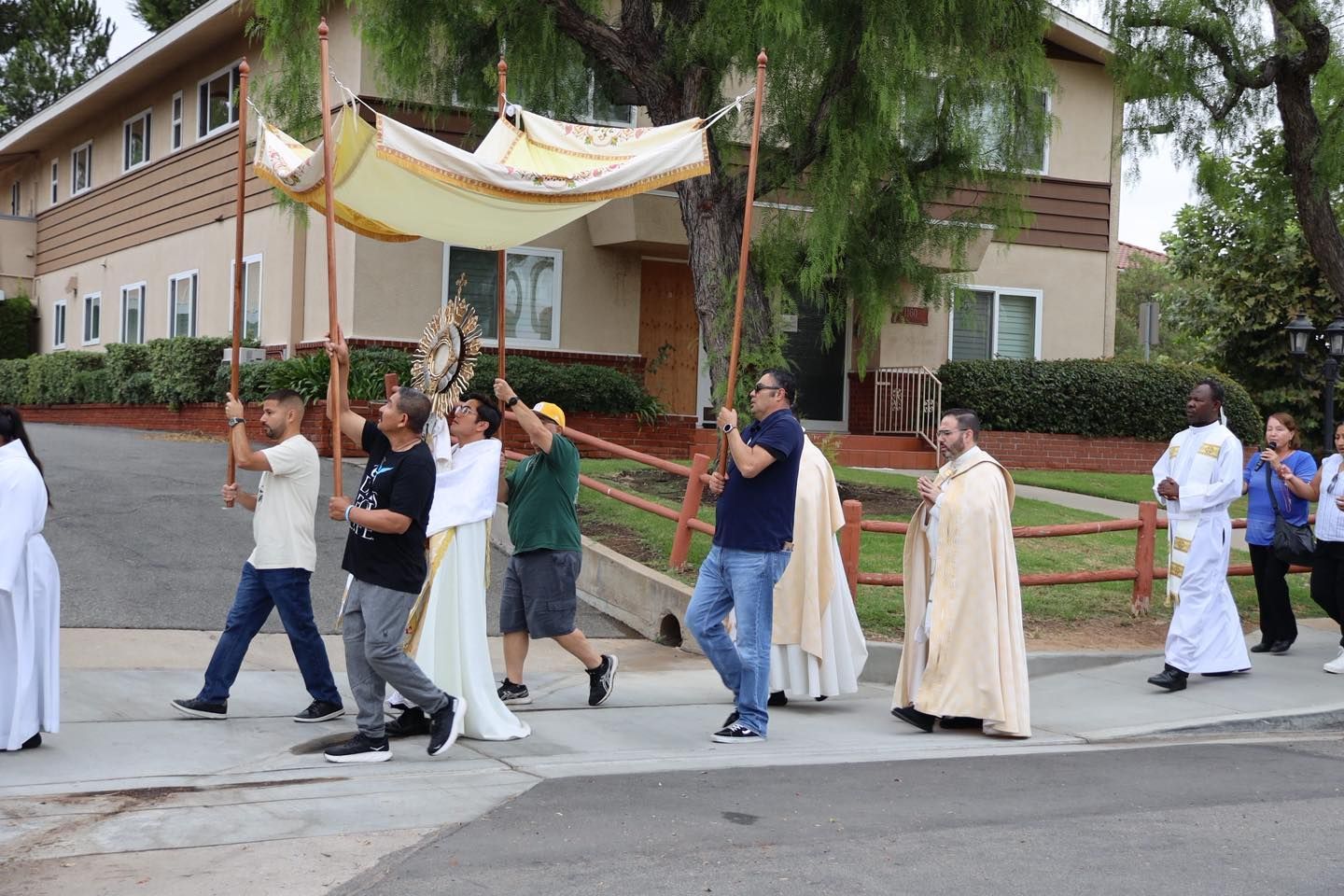 People carrying a canopy in a procession outside a building, with a priest walking beside them.