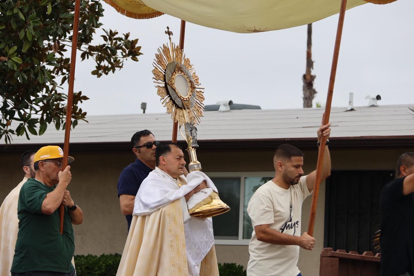 Clergy procession outdoors under a canopy, carrying a gold monstrance and a candlelit object
