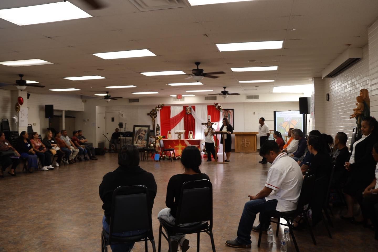 Indoor community gathering with people seated in a hall facing a small stage with red-and-white decorations