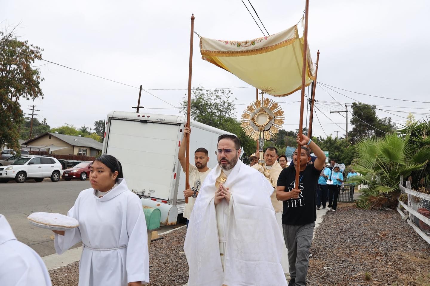 Religious procession outdoors, with people carrying a canopy and ornate cross on a gravel road.
