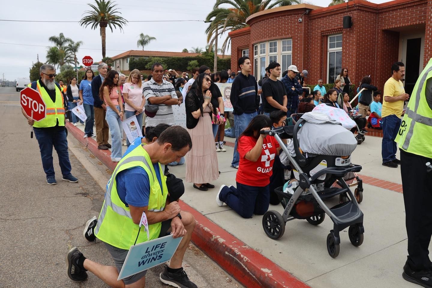 Protesters and volunteers gather on a street; one person holds a stop sign near a stroller and brick building.