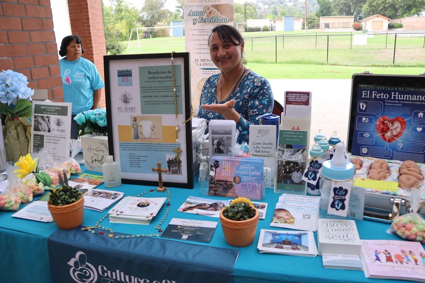 Community outreach table with brochures, flowers, jars, and two women standing behind it outdoors.