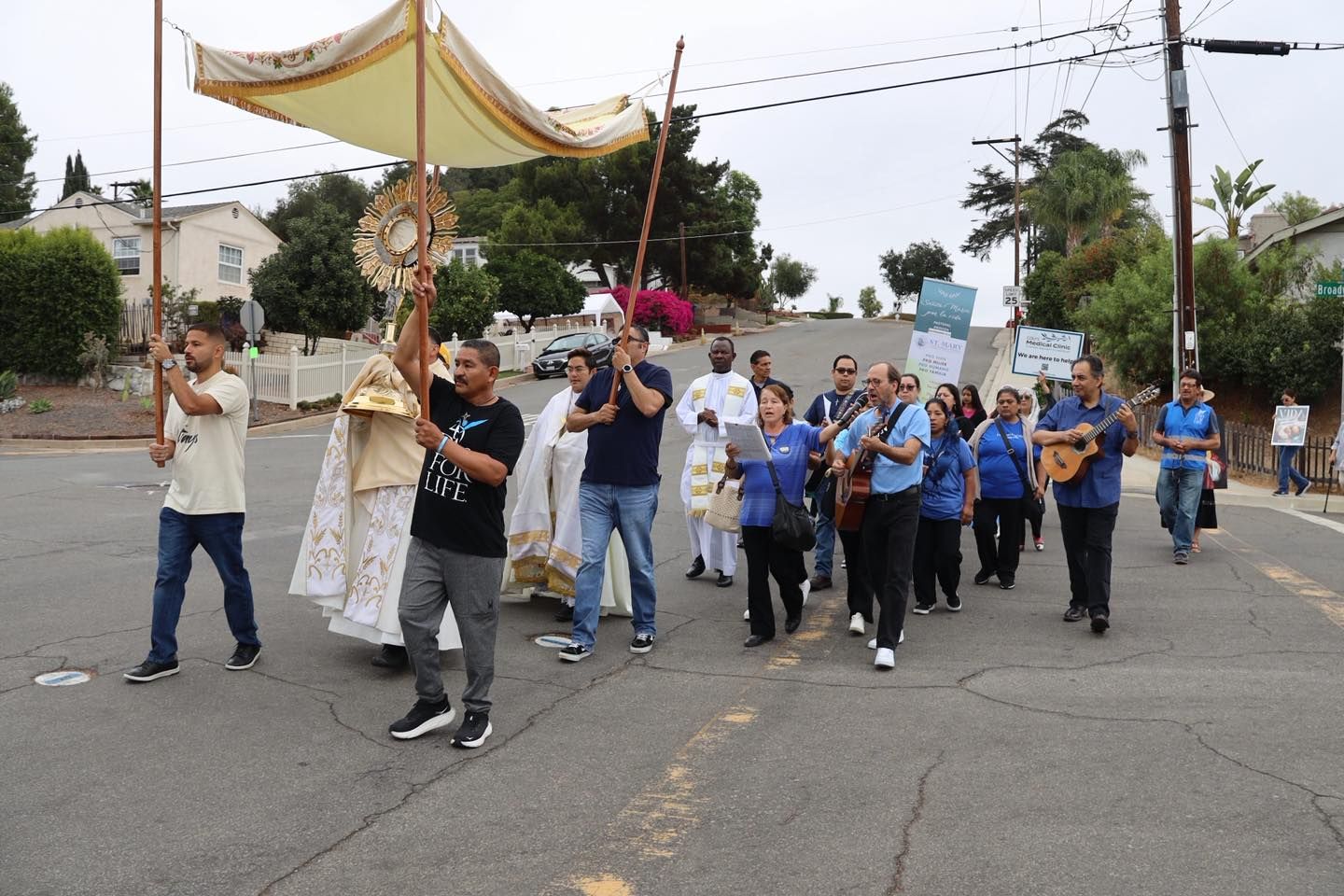 People walking in a street procession under a canopy, carrying a statue through a neighborhood.