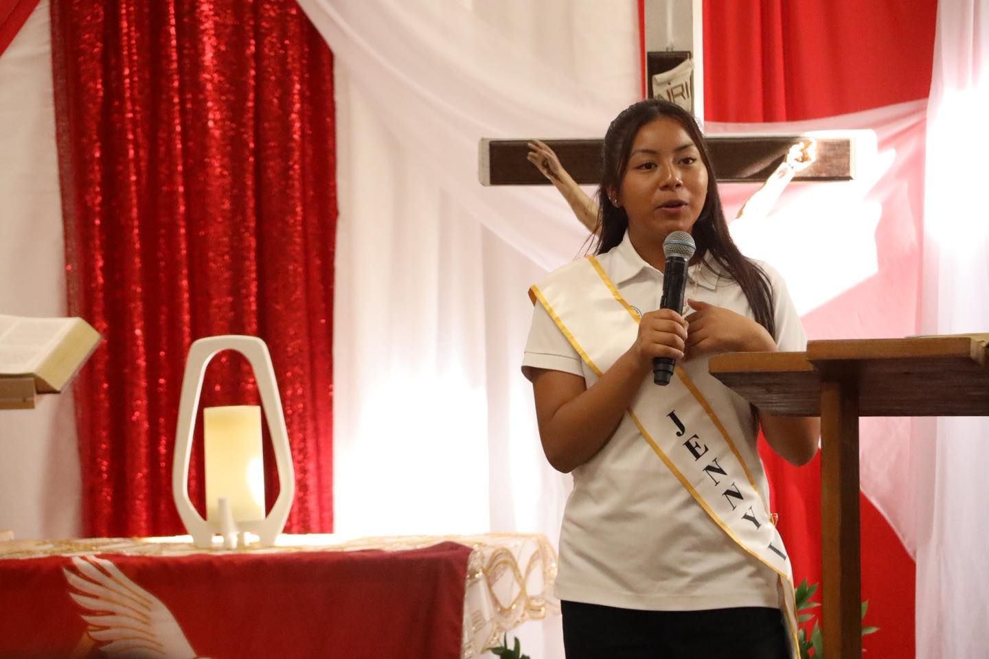 Woman speaking into a microphone at a red-and-white church altar with a cross behind her