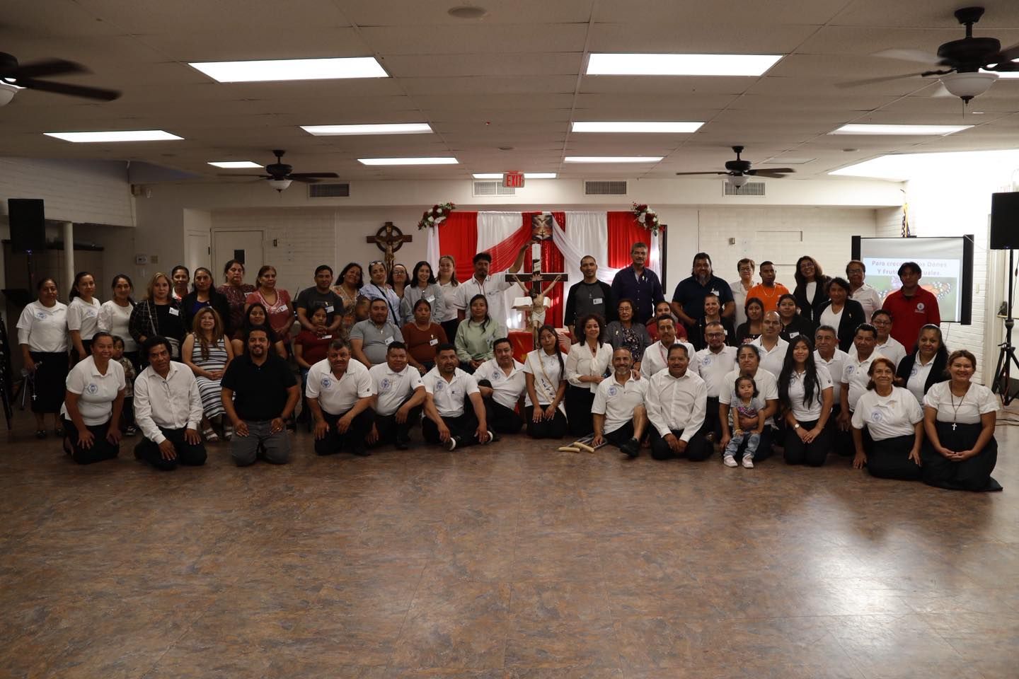 Large group posing in a bright hall, wearing mostly white shirts, with flags and red banners in the background.