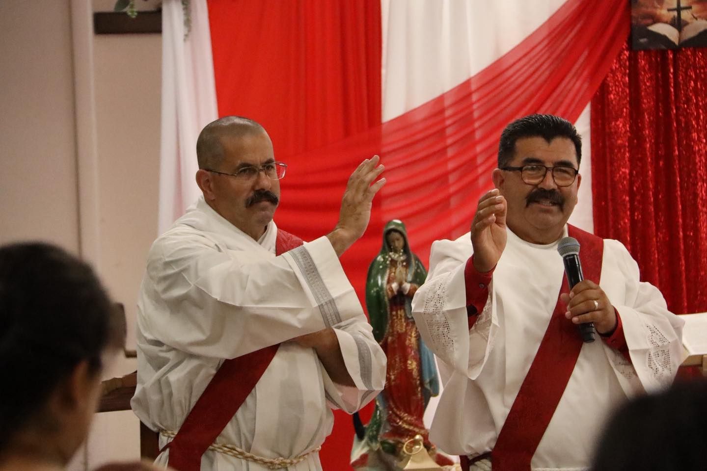 Two men in white robes with red sashes perform a traditional dance indoors with red-and-white drapes.