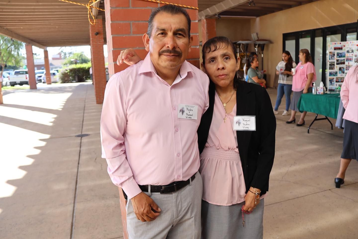 Two people standing together in a covered outdoor walkway at an event, one in a pink shirt and one in a black jacket.