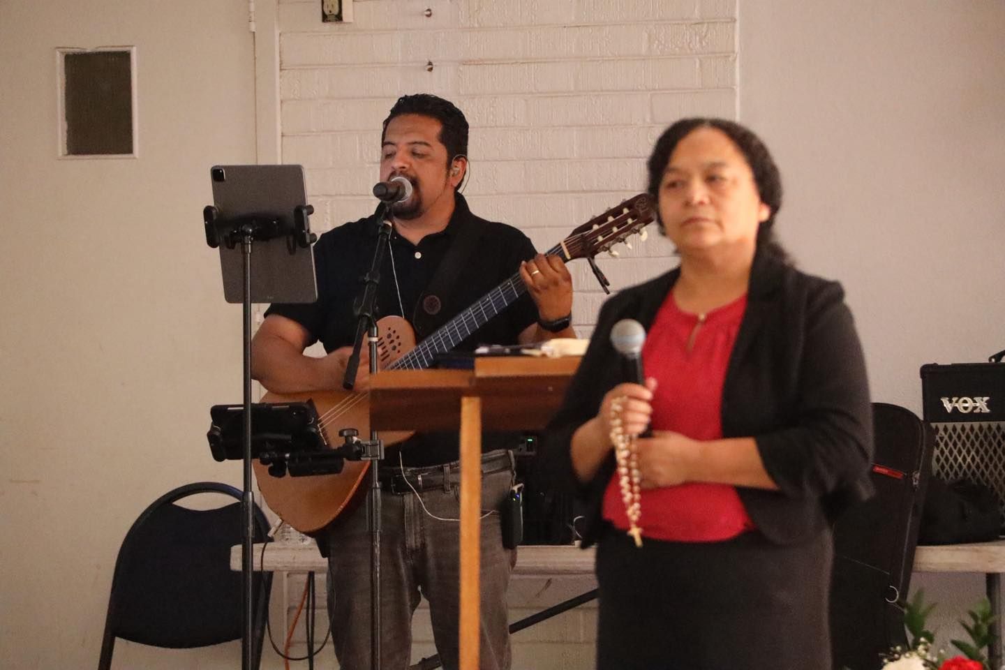 Two musicians performing indoors, with a singer holding a mic in front and a guitarist behind.