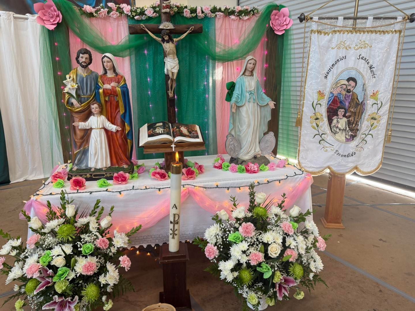 Religious altar display with crucifix, saints, flowers, candles, and pink-green drapery indoors