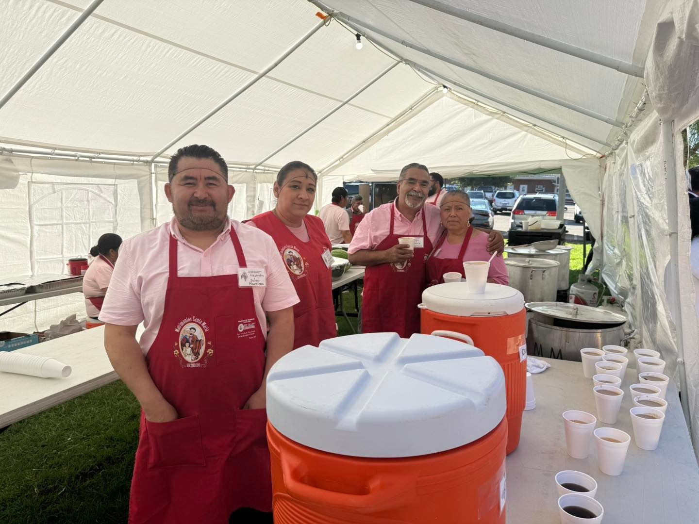 People in red aprons serving food and drinks under a white tent at an outdoor event
