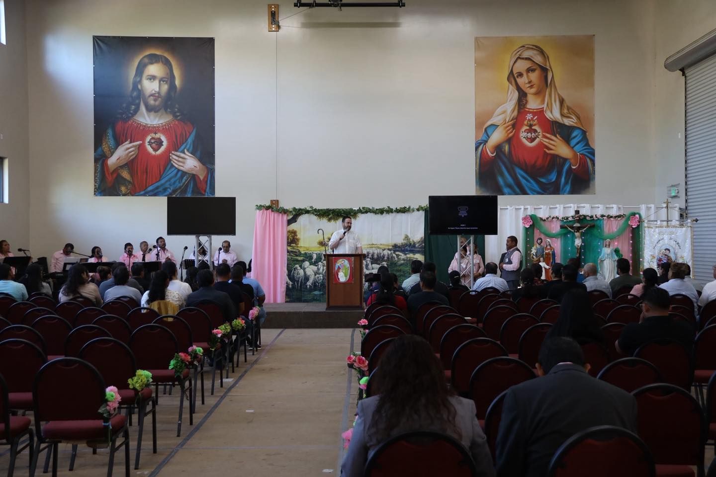 Indoor event hall with audience seated before a stage, podium, and large religious portraits on the wall