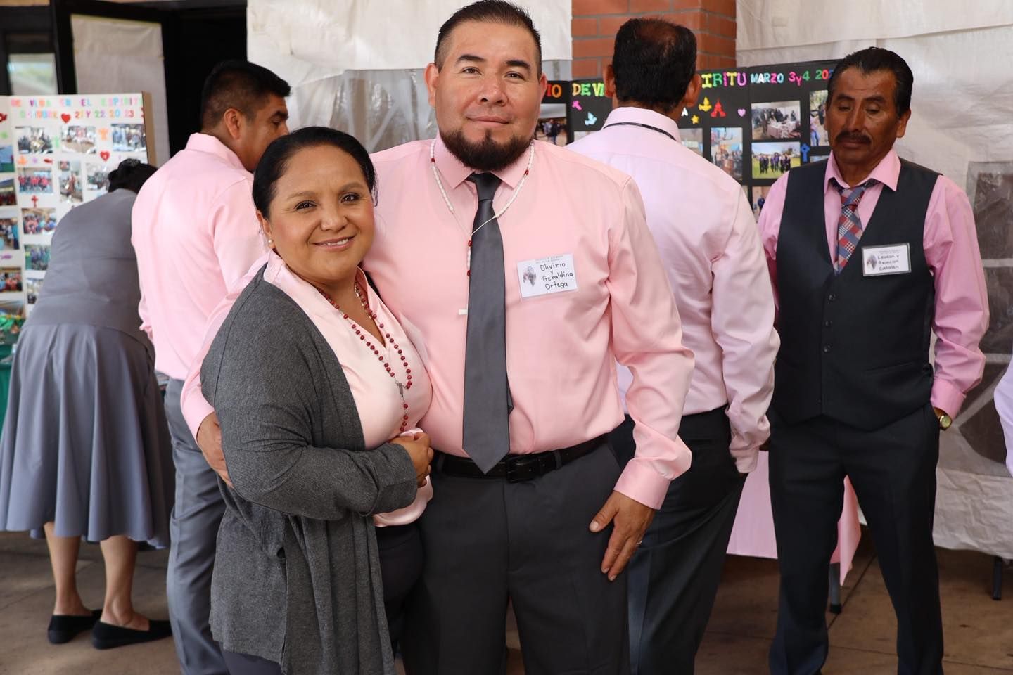 Two colleagues in pink shirts and gray attire pose smiling at an indoor event.
