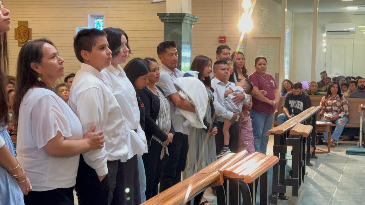 People standing in a church aisle during a ceremony, facing the front with benches nearby.