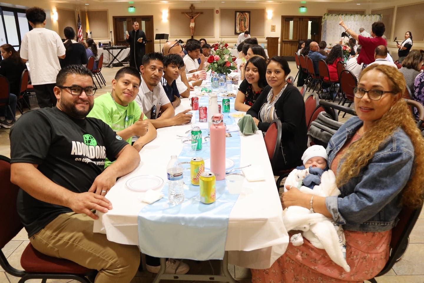 Group seated at a banquet table in a decorated hall, smiling with drinks and a baby in the foreground.