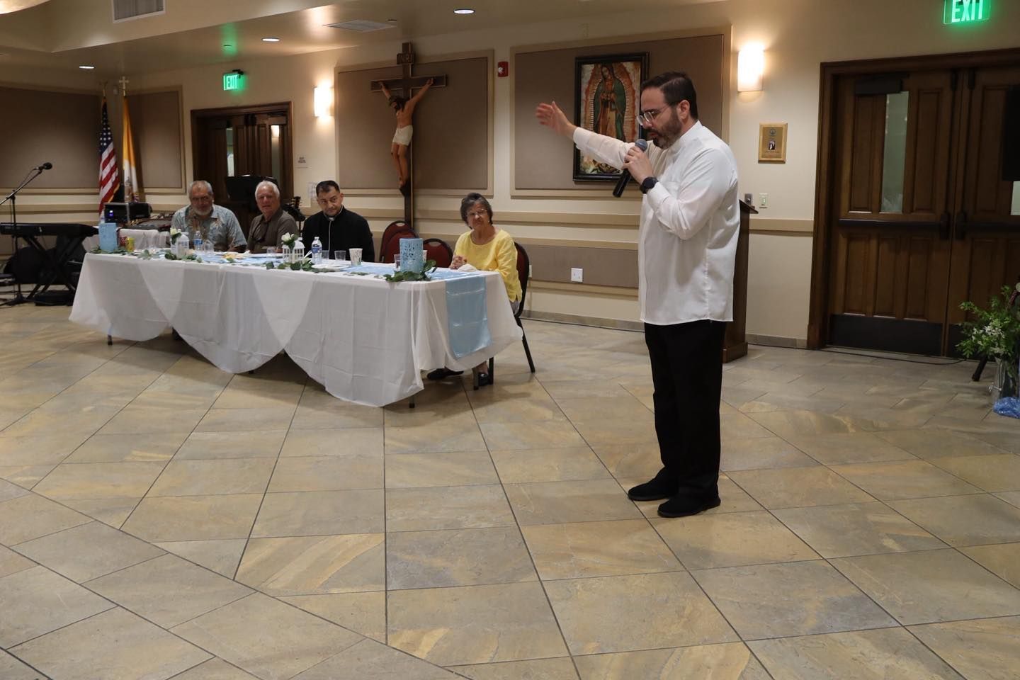 Man speaking at a banquet hall podium while seated panel listens at a long table