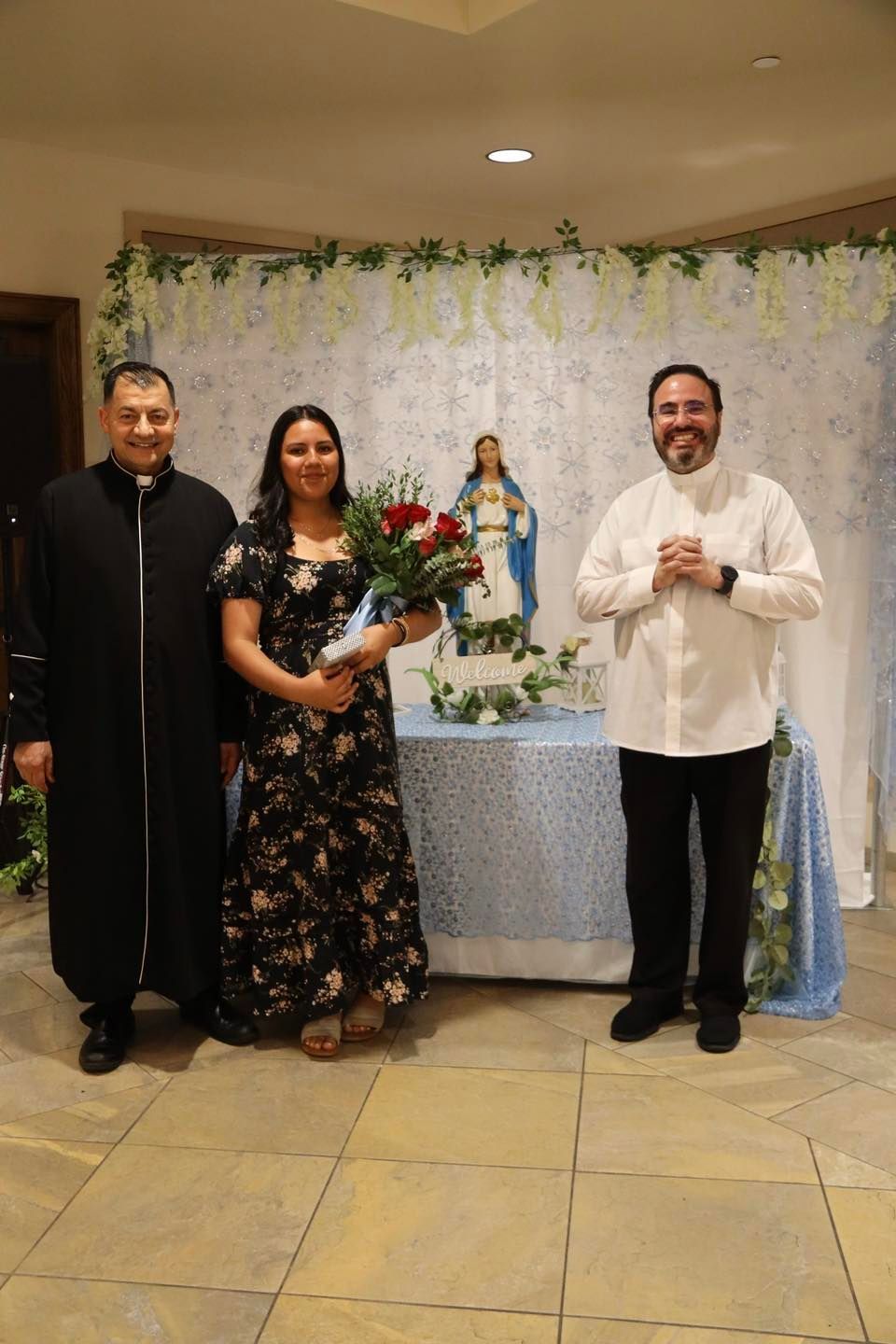 Three people posing beside a decorated religious altar with a statue and flowers.