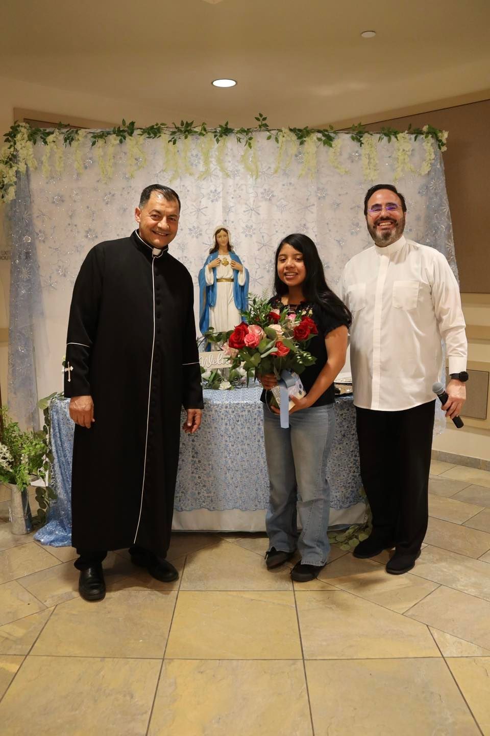 Three people pose indoors before a white floral backdrop and statue, with one holding a bouquet.