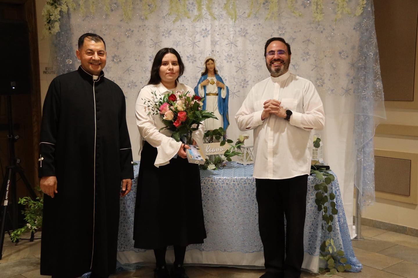 Three people standing before a small altar with a statue of Mary; center woman holds a bouquet.
