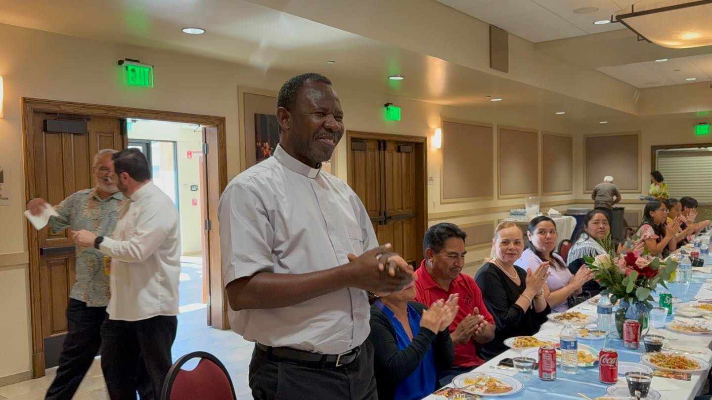 Man speaking beside a long banquet table at a community gathering, with people seated and applauding.