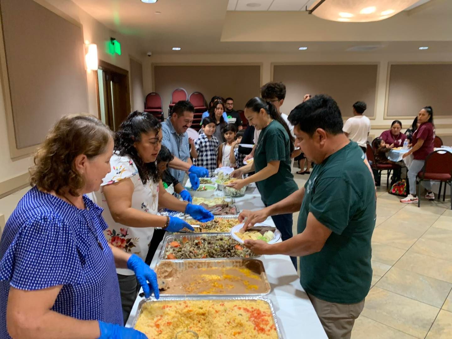 People serving food buffet-style in a banquet hall, with diners lining up and reaching for dishes.
