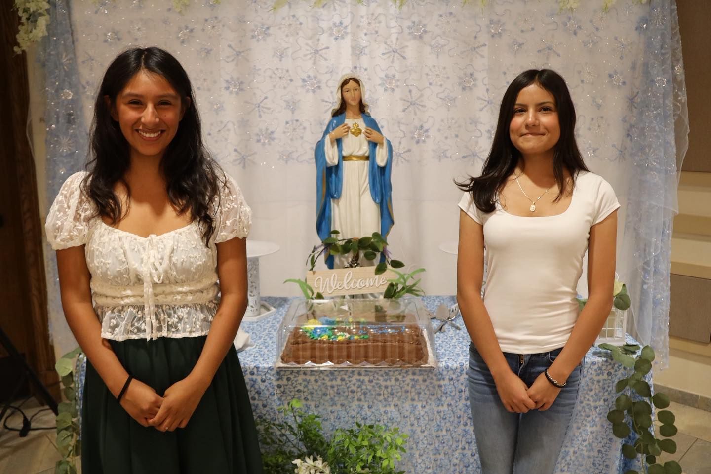 Two people standing beside a blue-and-white Virgin Mary altar with flowers and candles