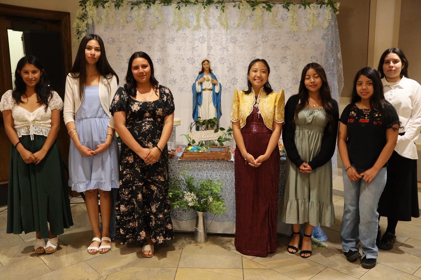 Seven people standing indoors before a decorated shrine and white floral backdrop, posing for a group photo