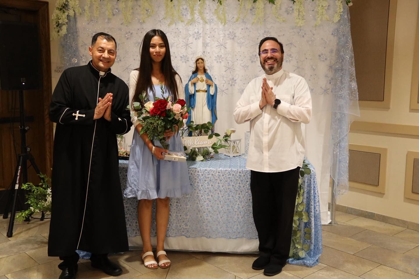 Three people standing beside a decorated altar with a statue, smiling and posing in a church hall