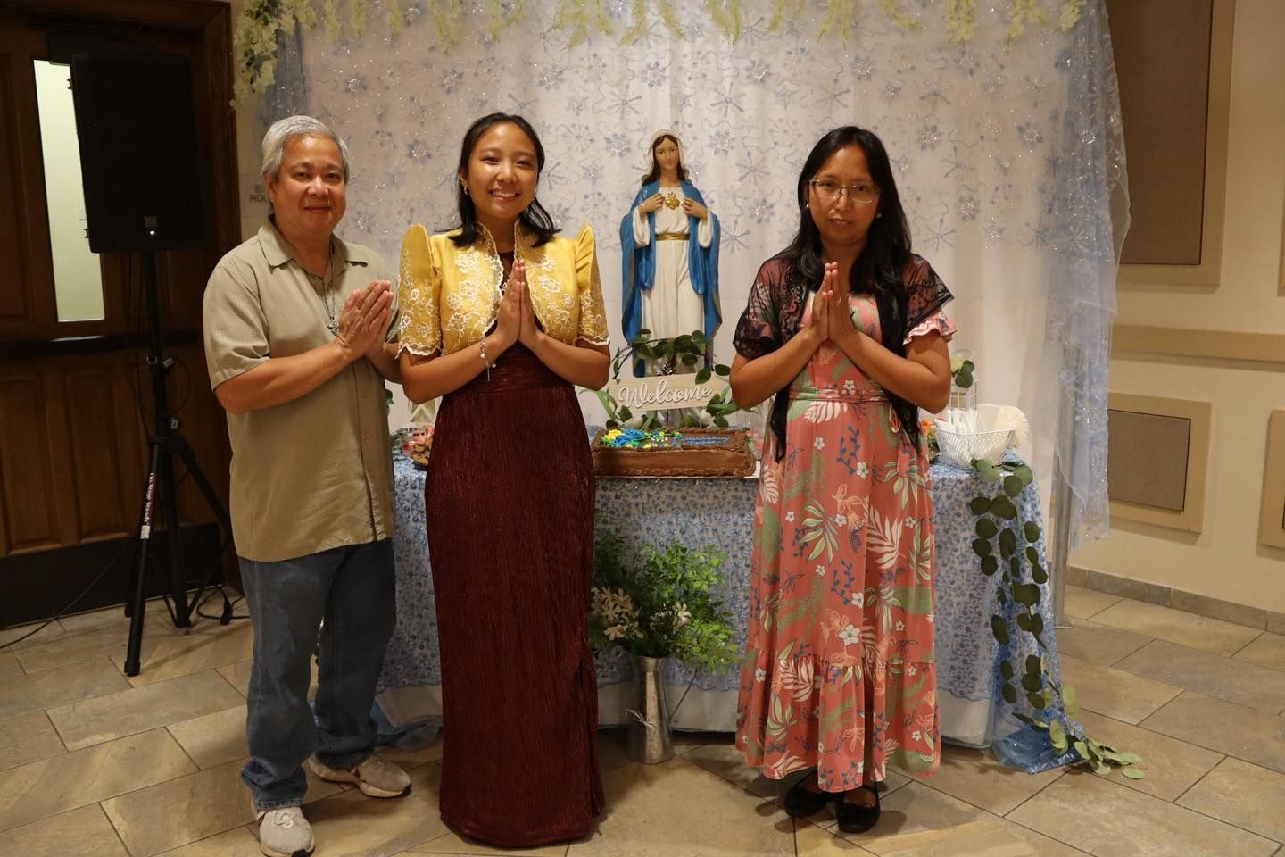 Three people stand with hands clasped before a shrine with a statue of the Virgin Mary.