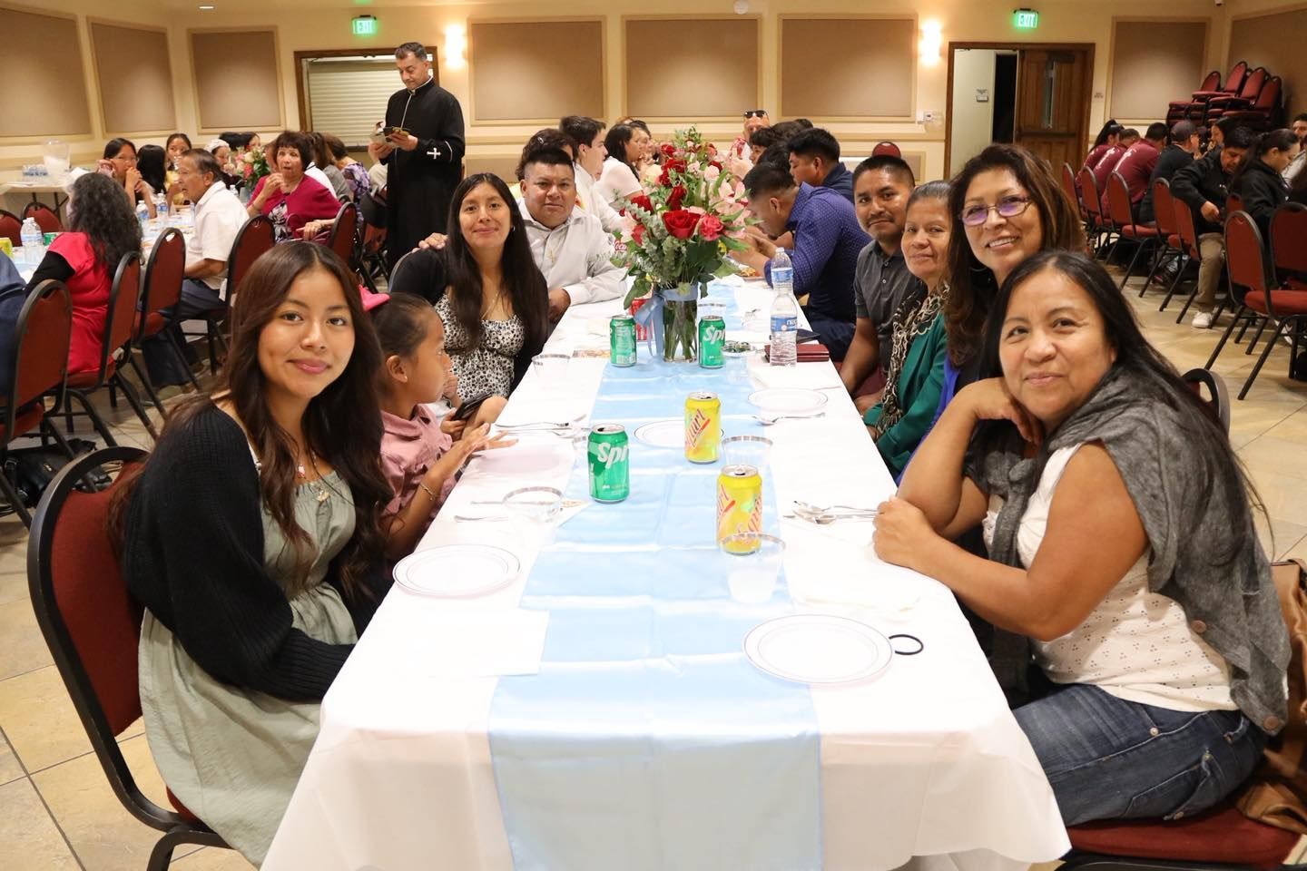 Long banquet table with seated guests, drinks, and a floral centerpiece in a hall.