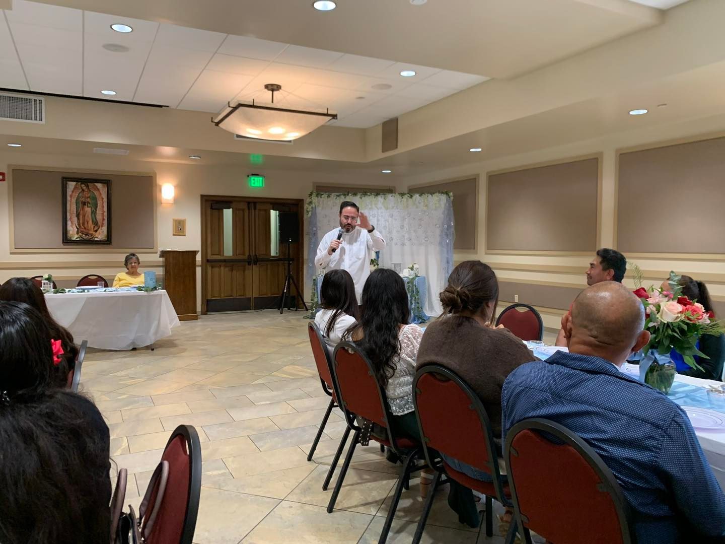 Man speaking at a microphone in a banquet hall, with seated guests facing him and floral centerpieces on tables.