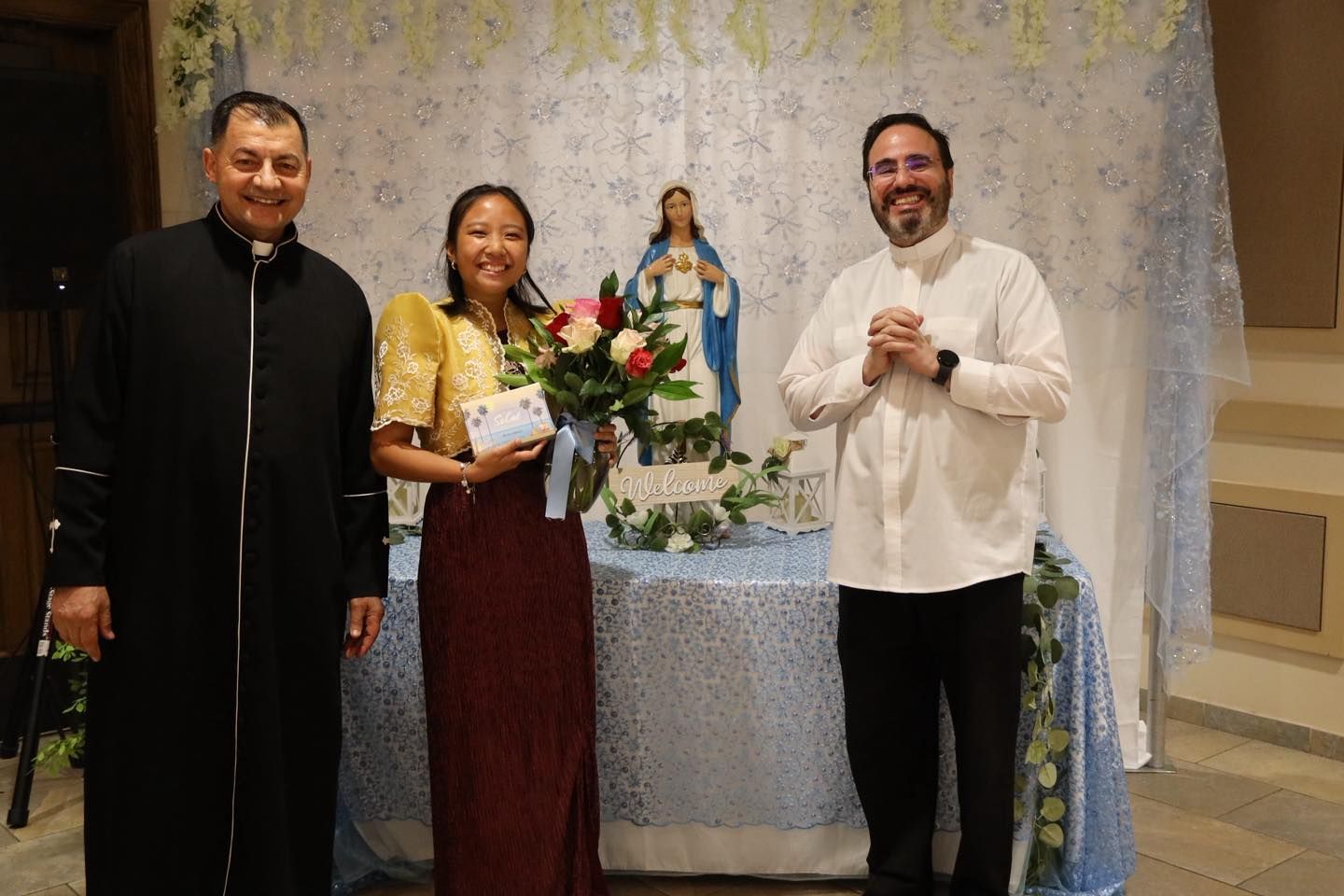 Three people posing indoors beside a flower-decorated table and statue at a formal event.