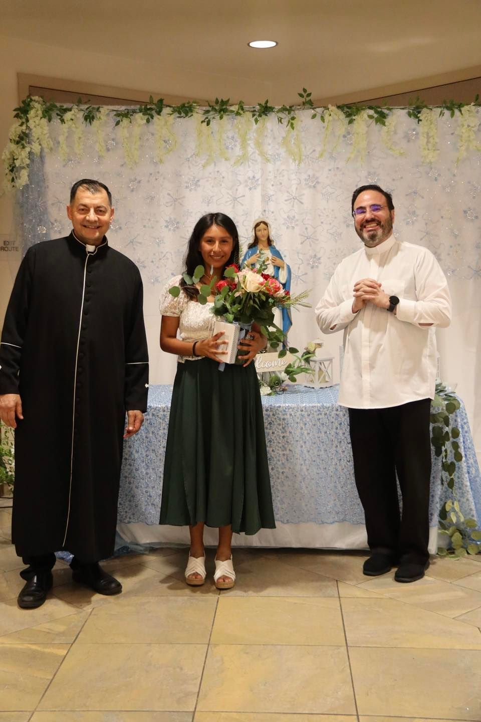 Three people standing indoors at a decorated event table, holding bouquets and smiling.