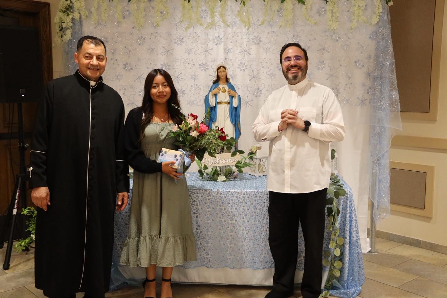 Three people standing indoors beside a flower-filled table and a statue of the Virgin Mary