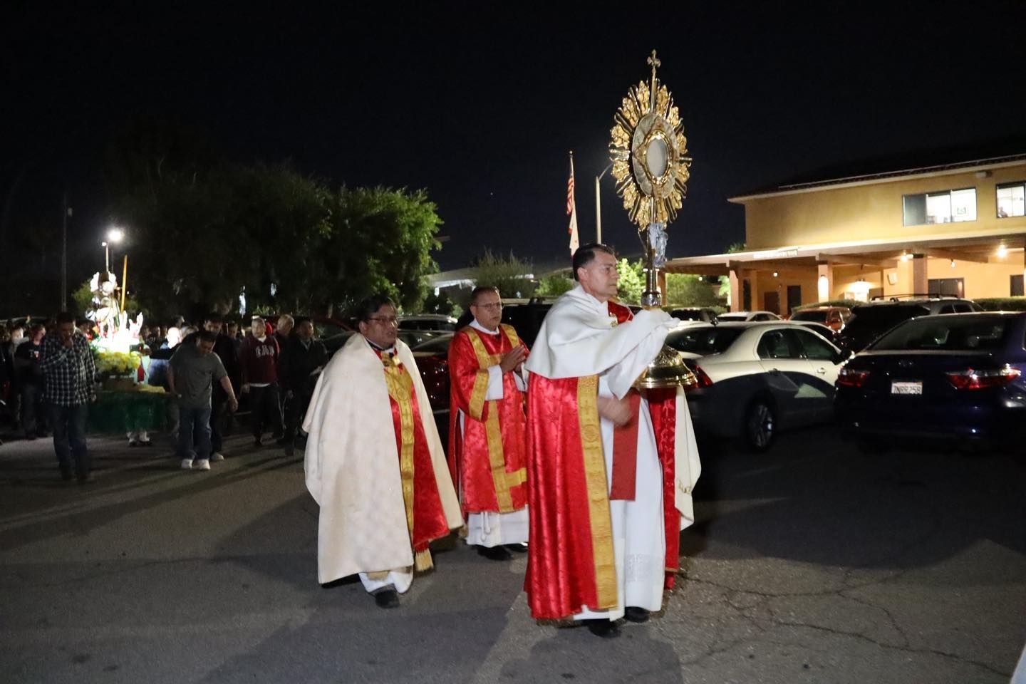 Clergy in white and red robes carry a decorated religious banner during a nighttime procession.