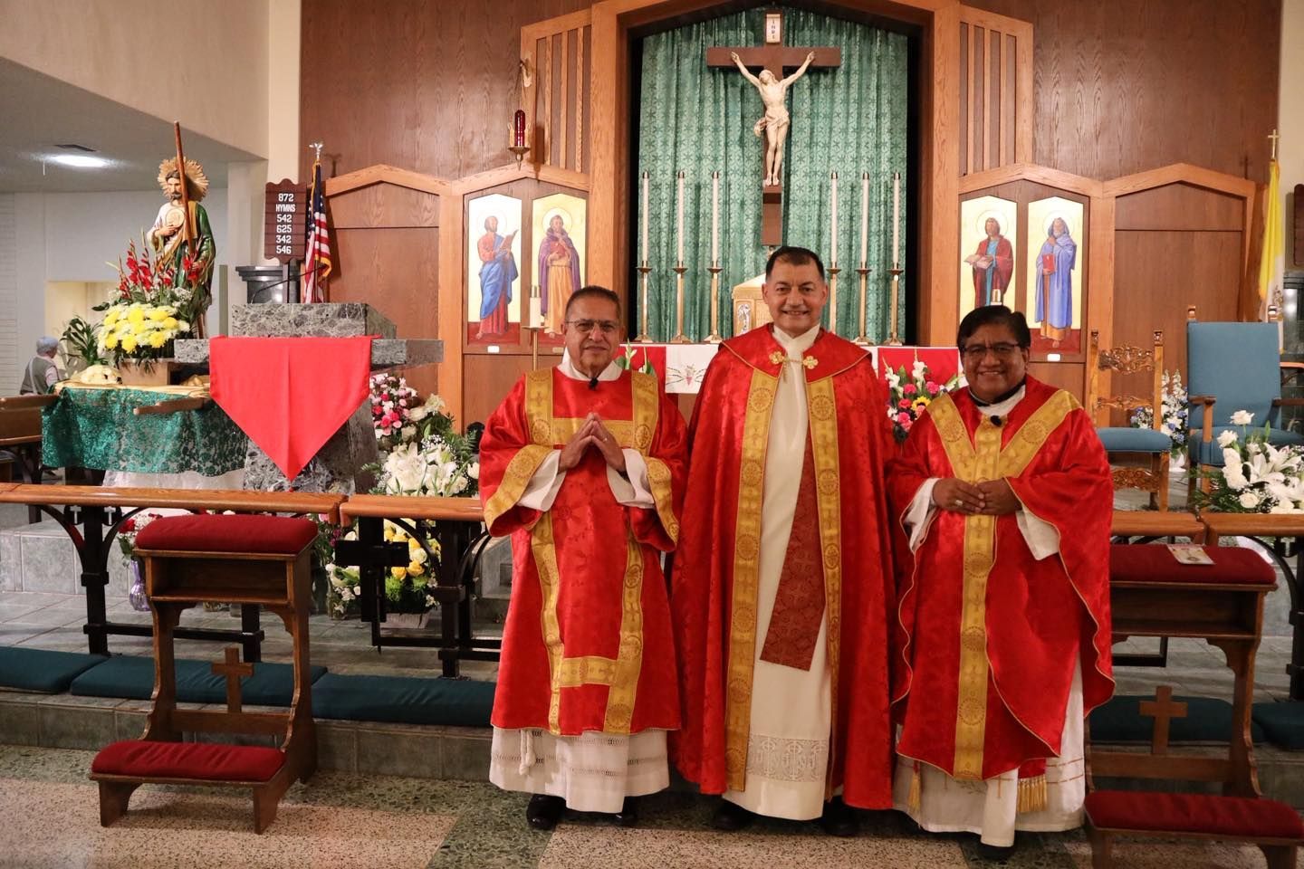 Three clergy in red vestments stand before a church altar and crucifix.