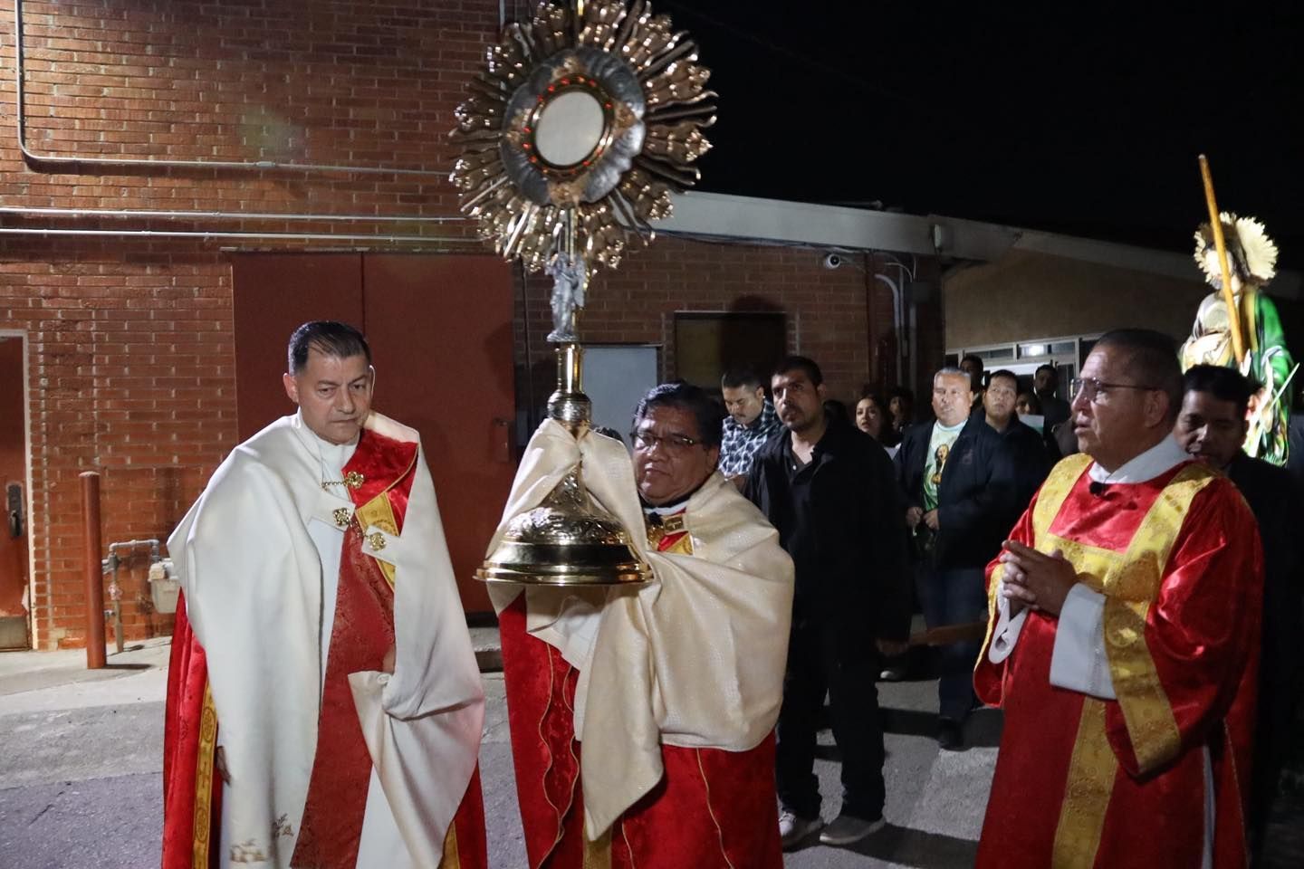 Clergy in white and red robes carrying a gold monstrance and bell in a church procession