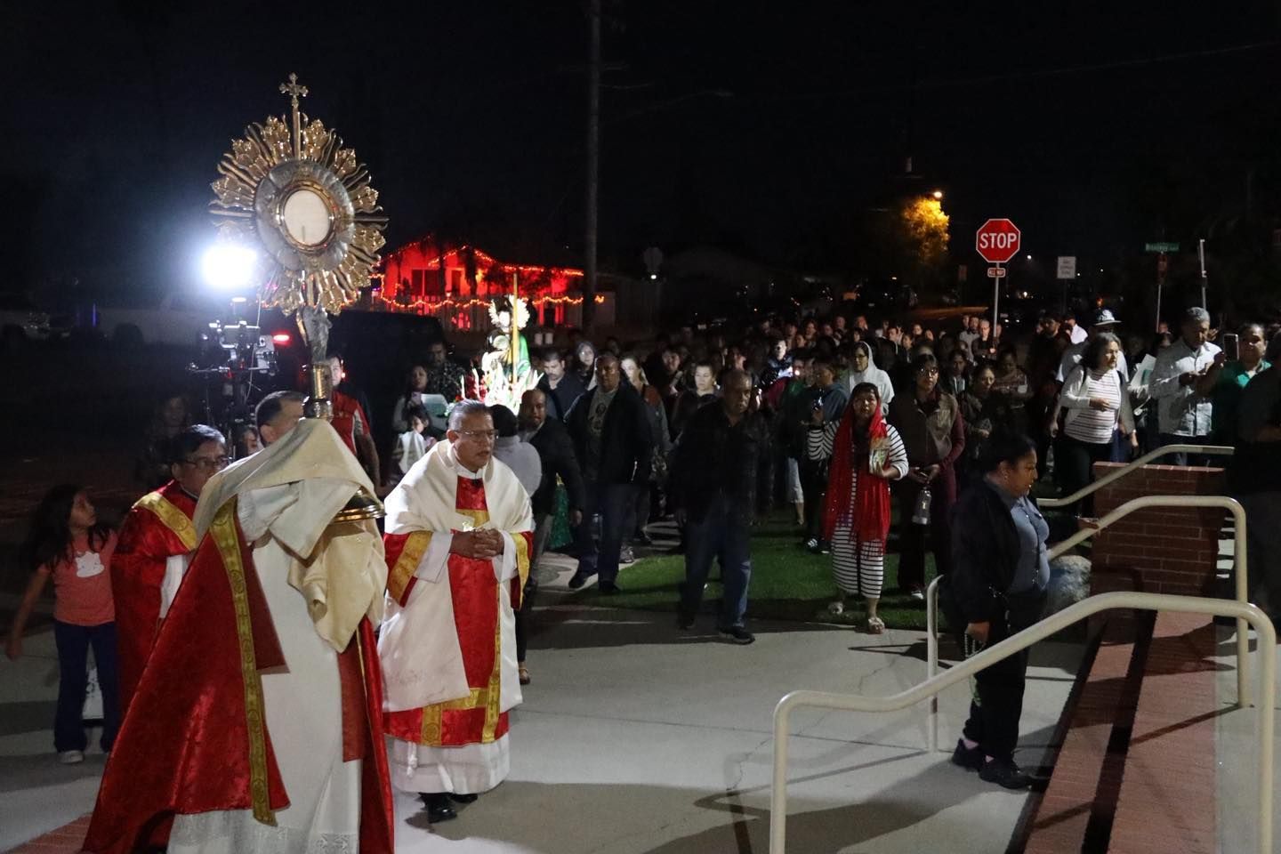 Clergy in white and red robes lead a nighttime outdoor religious procession before a crowd