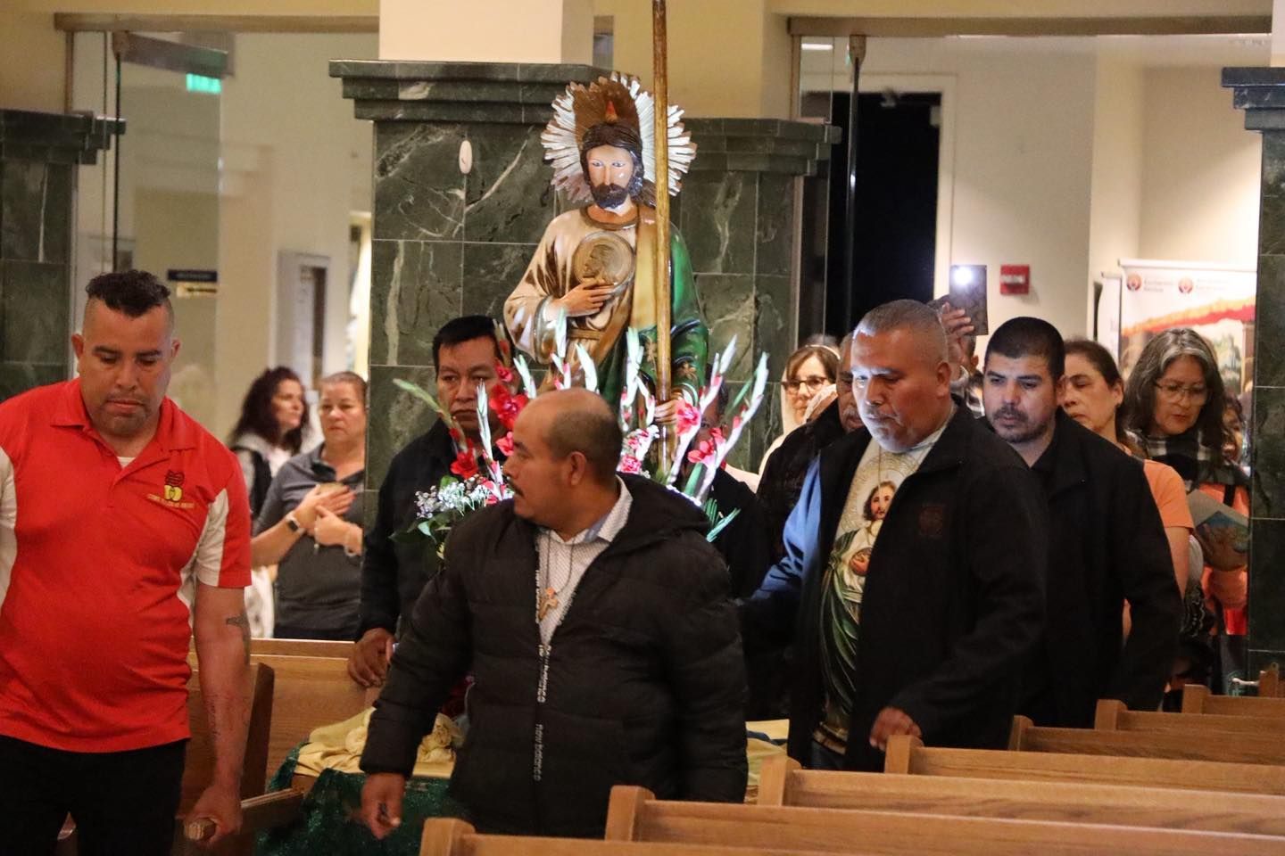People seated in church pews during a procession, with a statue carried down the aisle.