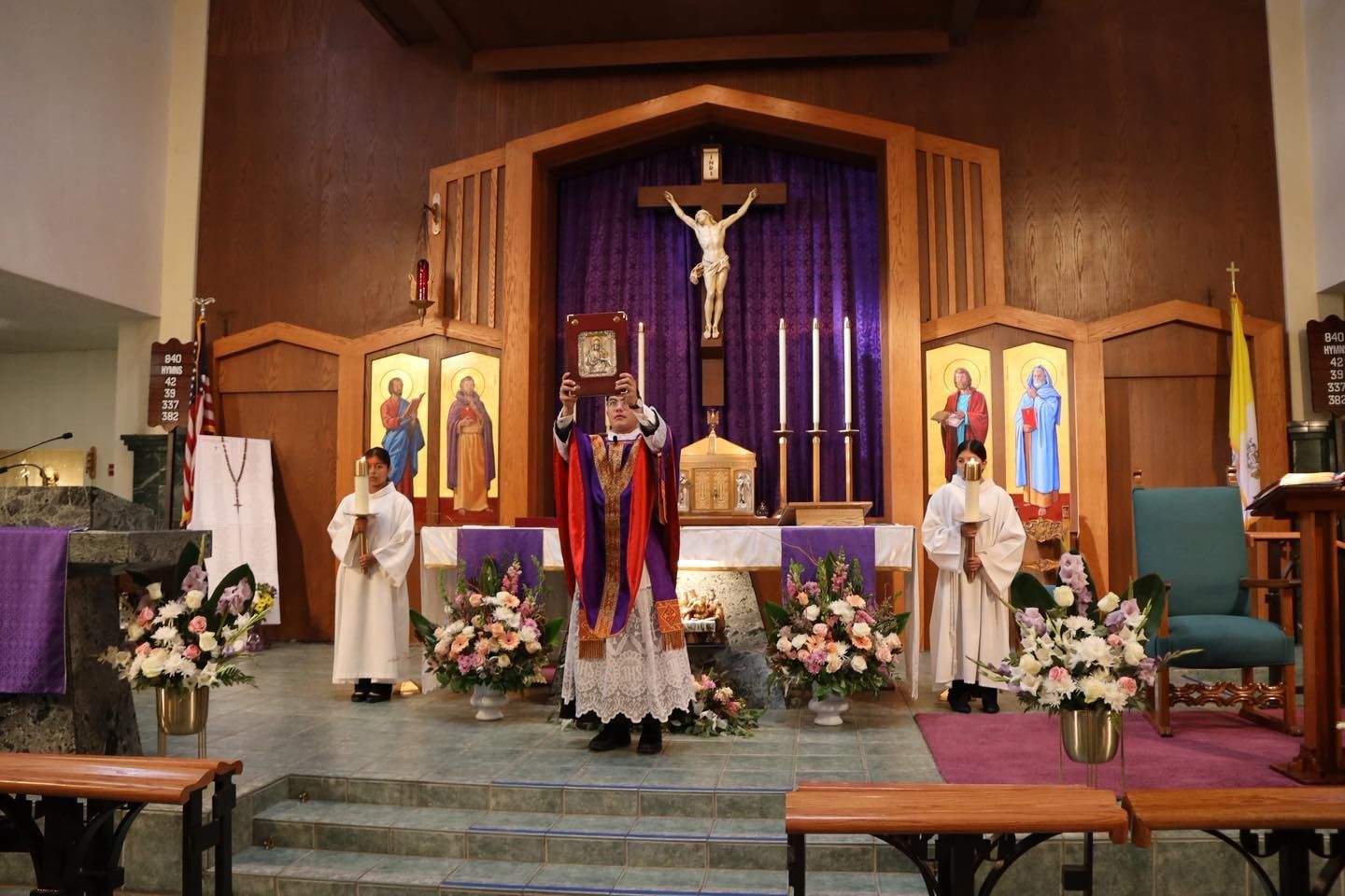 Religious altar scene with a crucifix, statues, flowers, and people in white robes standing in a church.