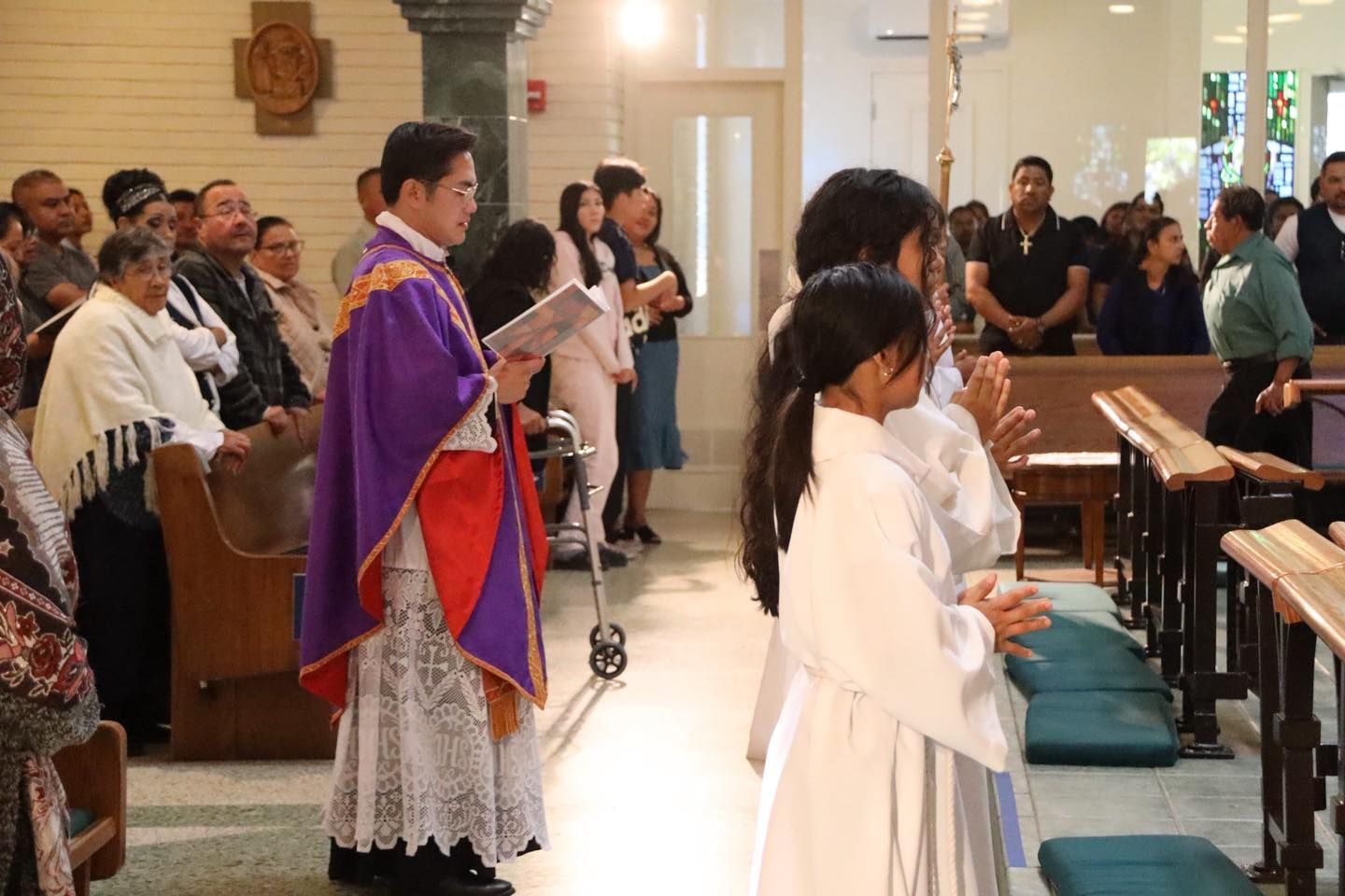 Priest in purple vestments leads a church service with altar servers and congregants standing nearby