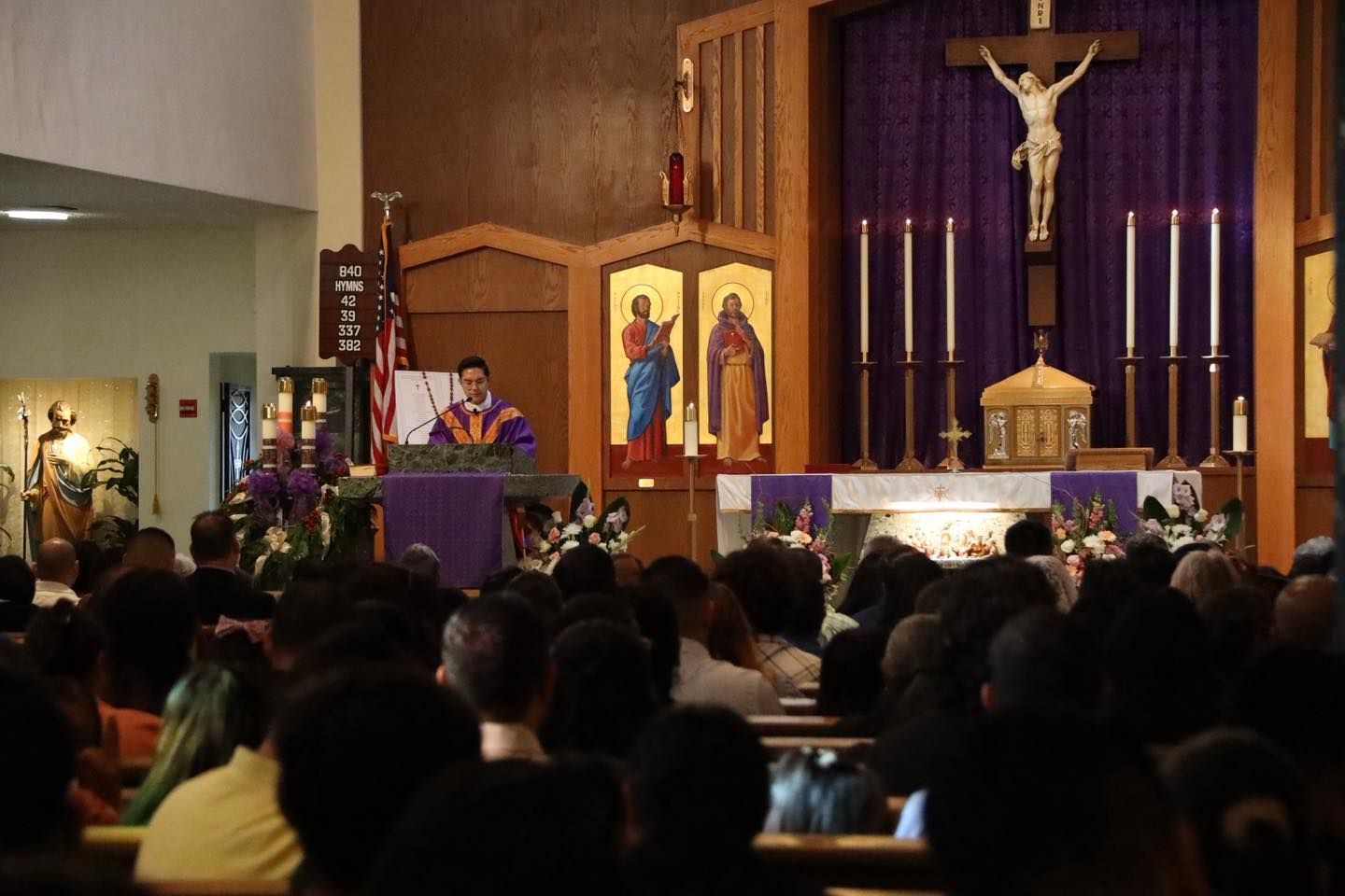 Church service with a priest at the altar beneath a crucifix, congregation seated in pews.