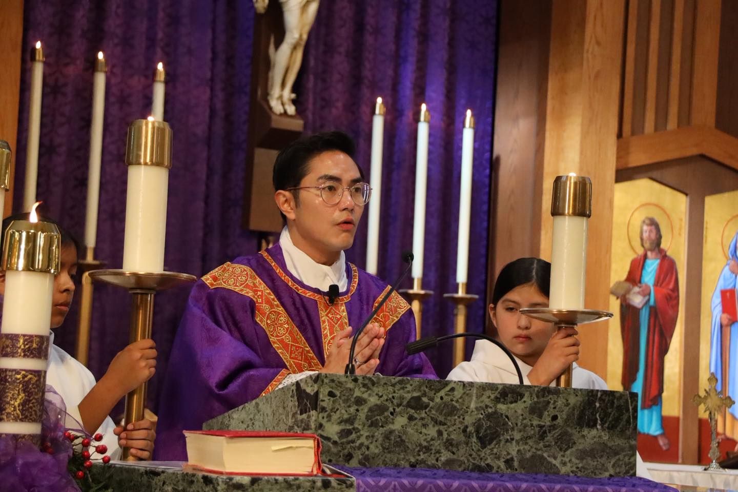Priest in purple vestments at a church altar during Mass, with candles and a server beside him
