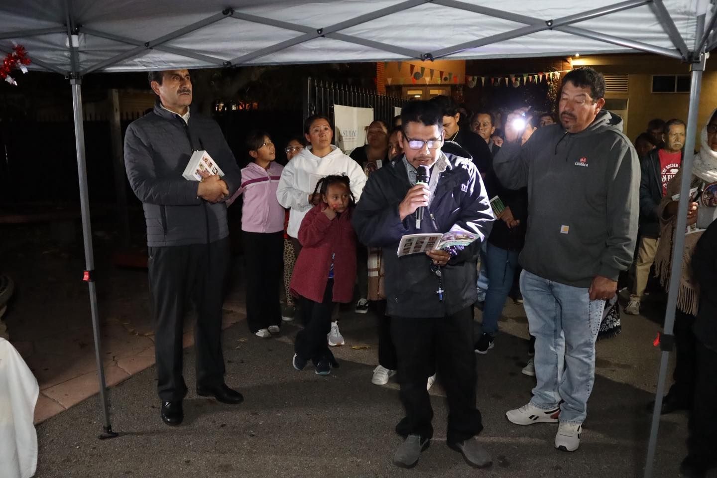 People standing under a canopy at night, one reading from a paper while others watch.