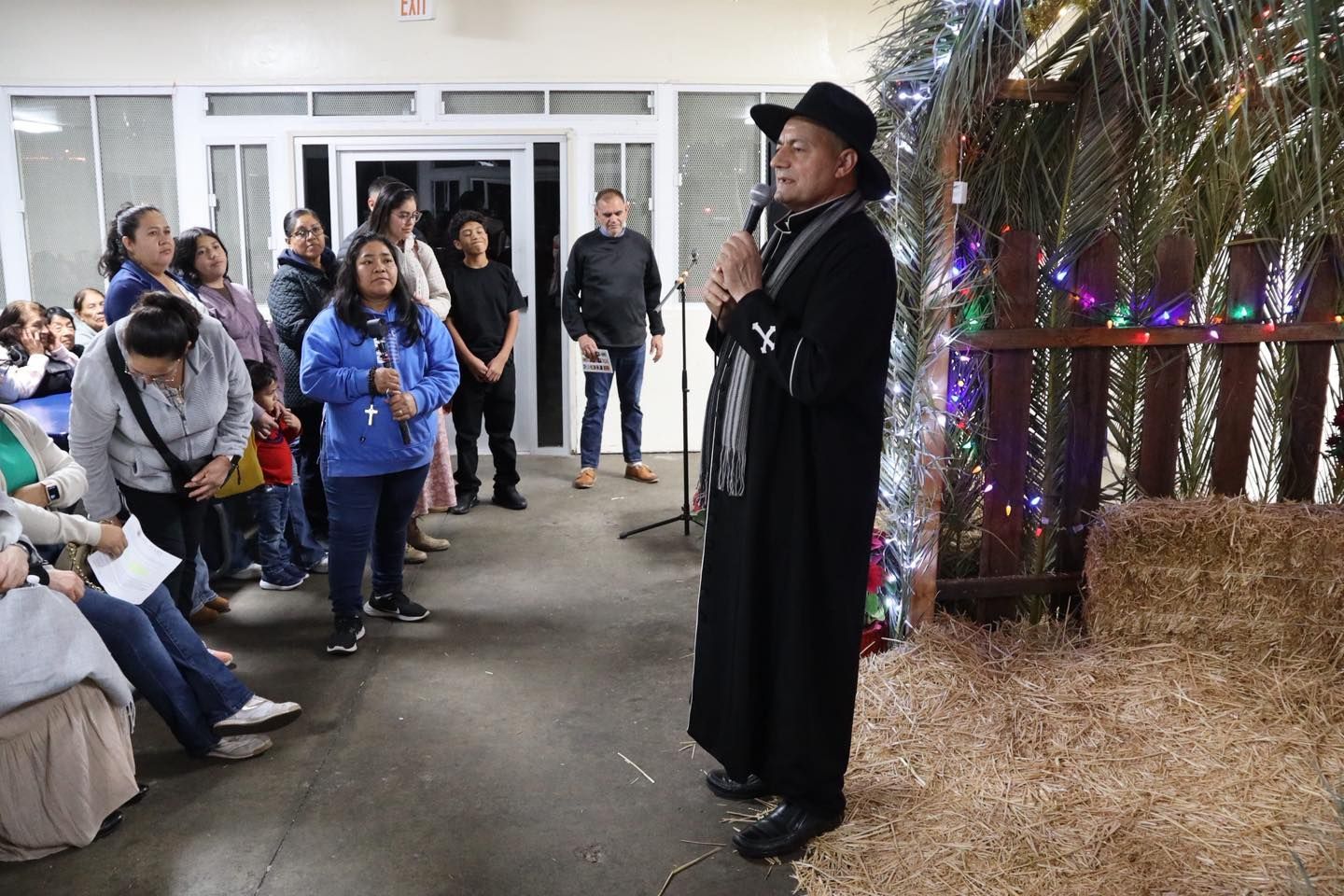 Man in black coat speaking to a seated crowd indoors beside a decorated, straw-lined display